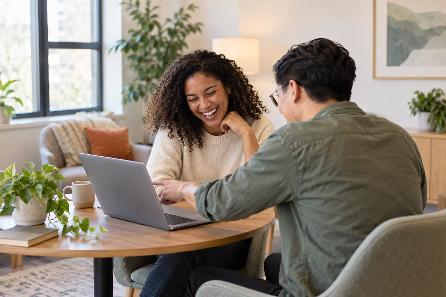 Two coworkers share one laptop at a round table, one pointing at the screen while the other laughs.