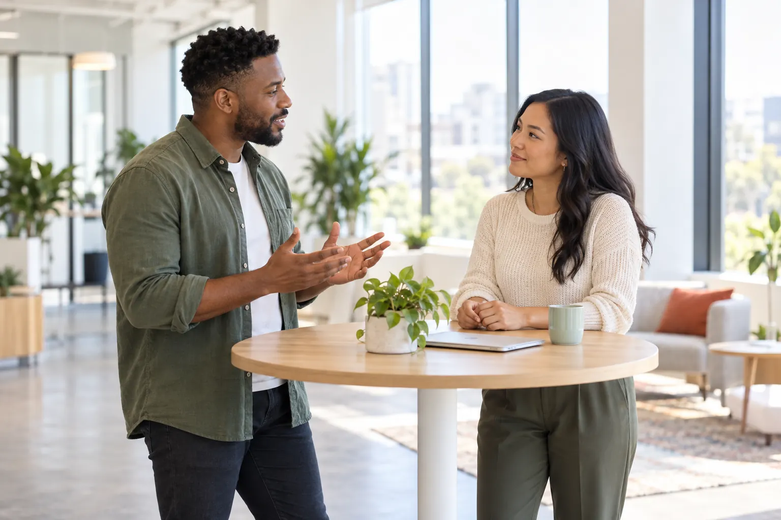 Two coworkers talk at a standing table in a bright office, one gesturing while the other listens.