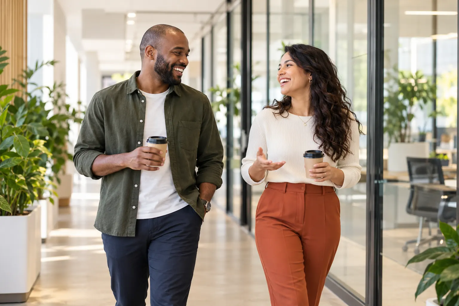 Two coworkers share a laugh over takeaway coffee as they walk past glass-walled offices.