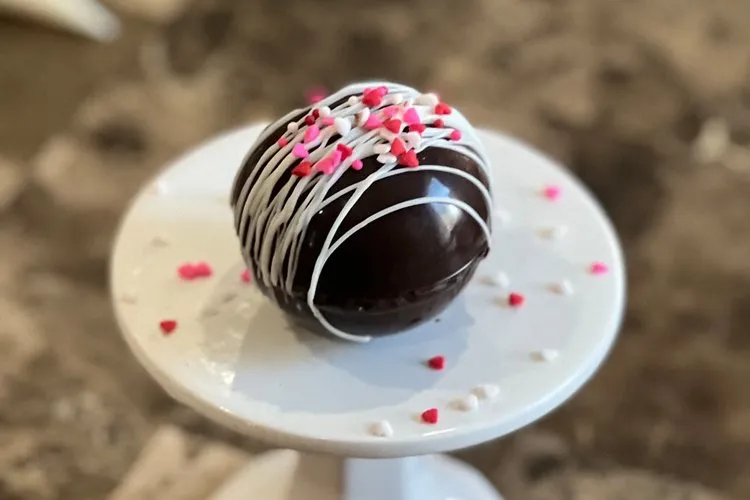 A finished hot chocolate bomb with white drizzle and heart sprinkles