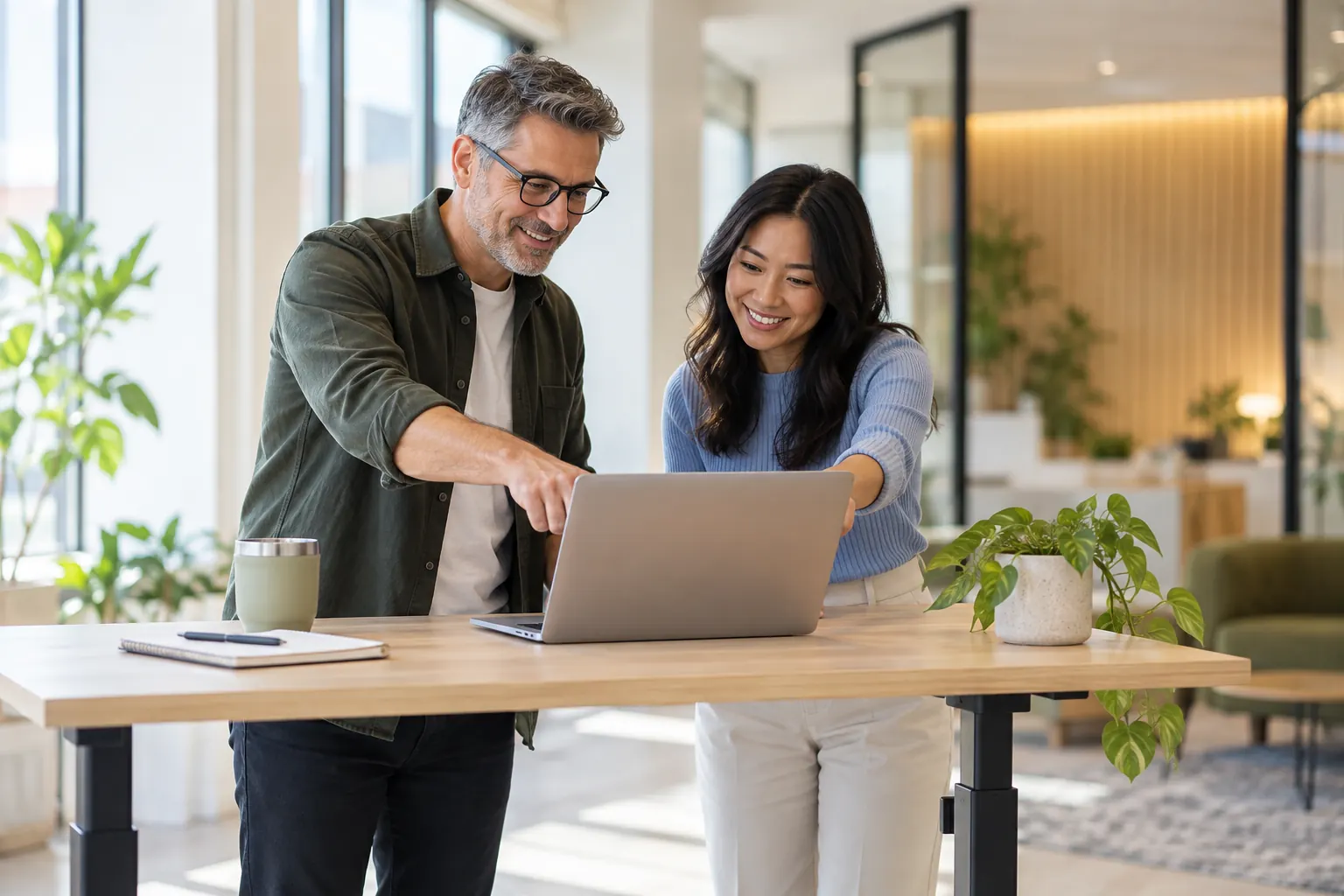 Two colleagues of different ages smile at a laptop on a standing desk in a bright office.