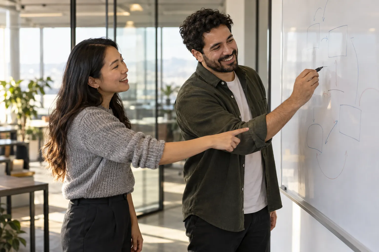 Two coworkers smile at a whiteboard, one sketching boxes and arrows while the other points at the drawing