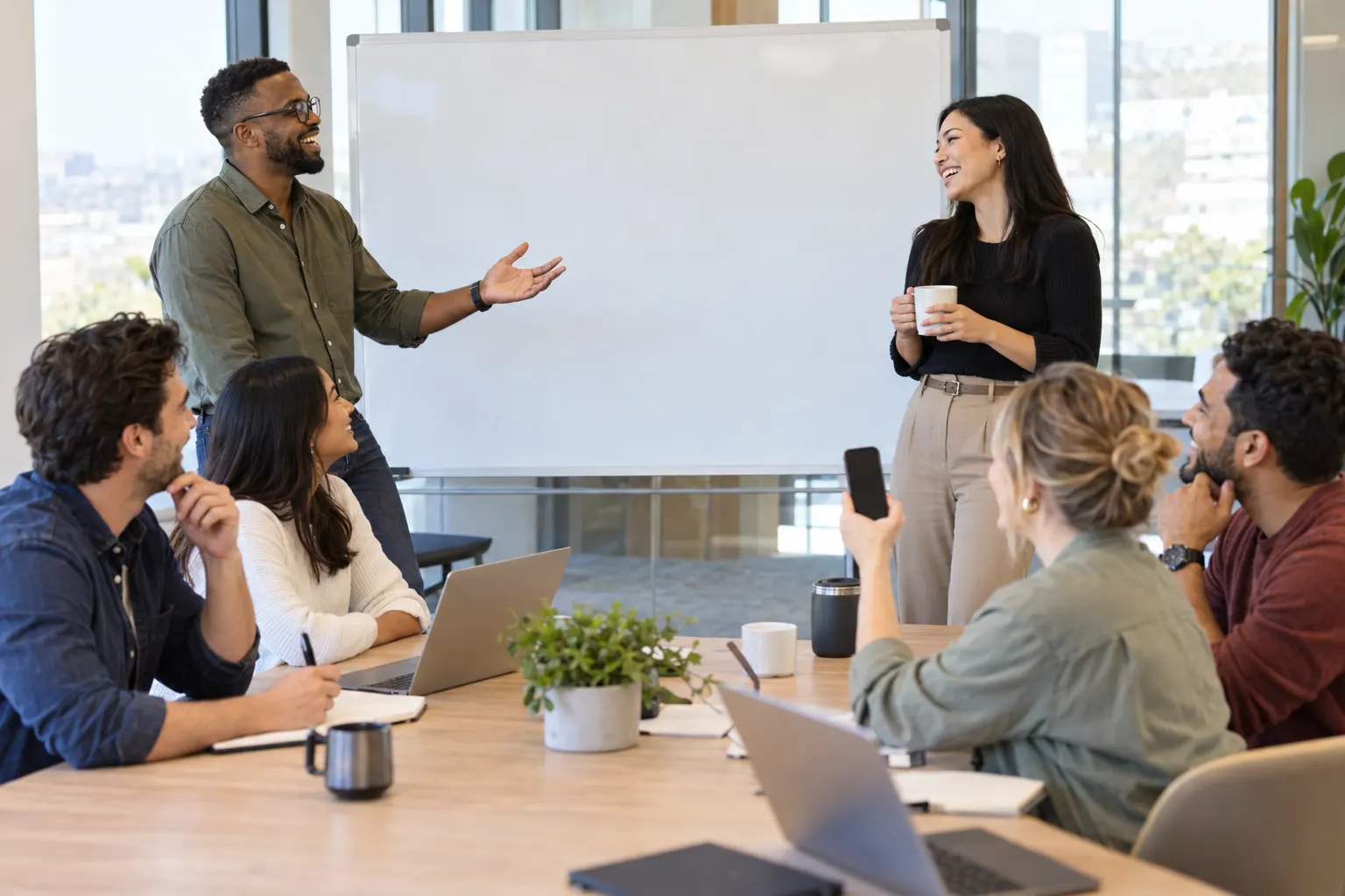 Two coworkers trade a playful argument by a whiteboard while seated colleagues laugh and one holds up a phone