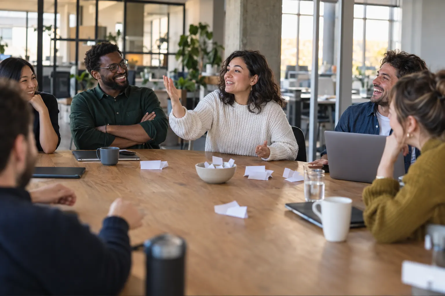 A coworker makes a playful argument at an office table while teammates laugh, with folded paper slips in a bowl between them