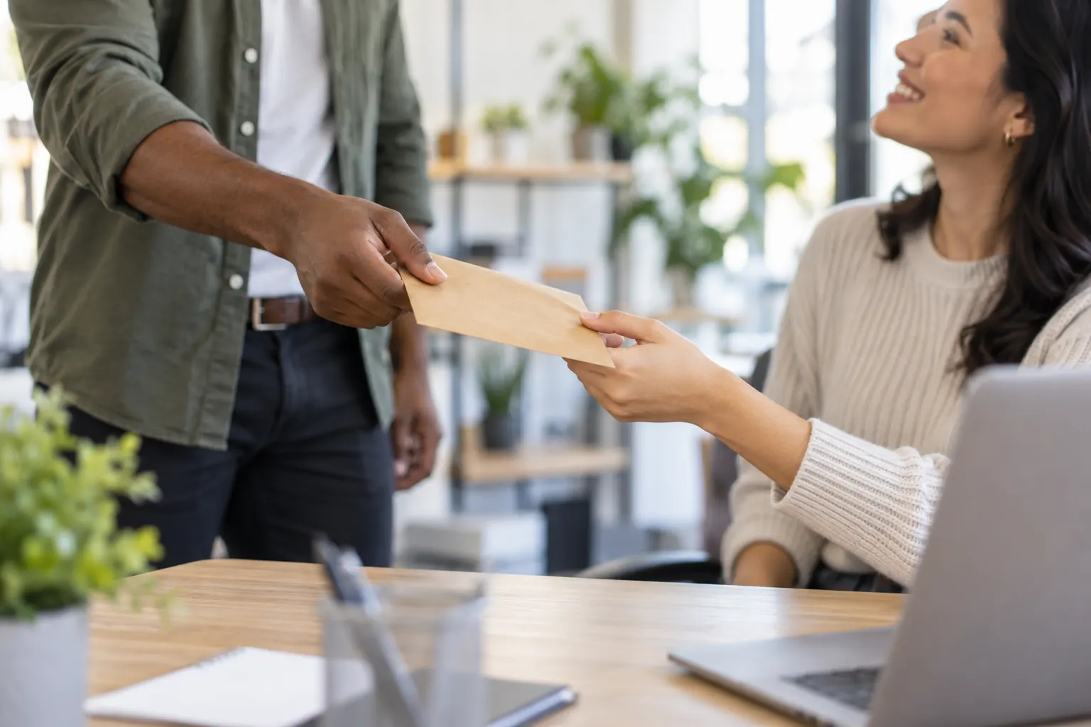 A manager hands a small kraft envelope to a smiling employee sitting at her desk.