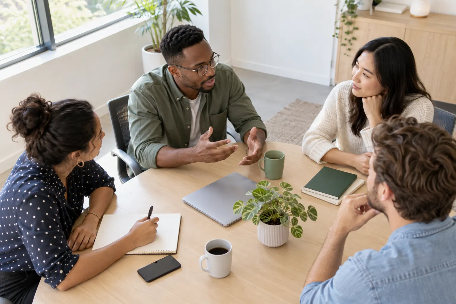 Seen from above, four colleagues at a meeting table: one leans in to speak, two listen, one takes notes.