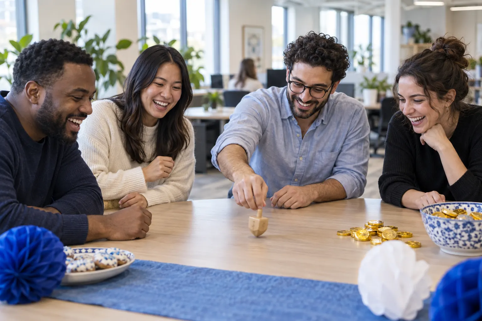 Four coworkers laughing around a table as one spins a wooden dreidel beside a pile of chocolate coins.