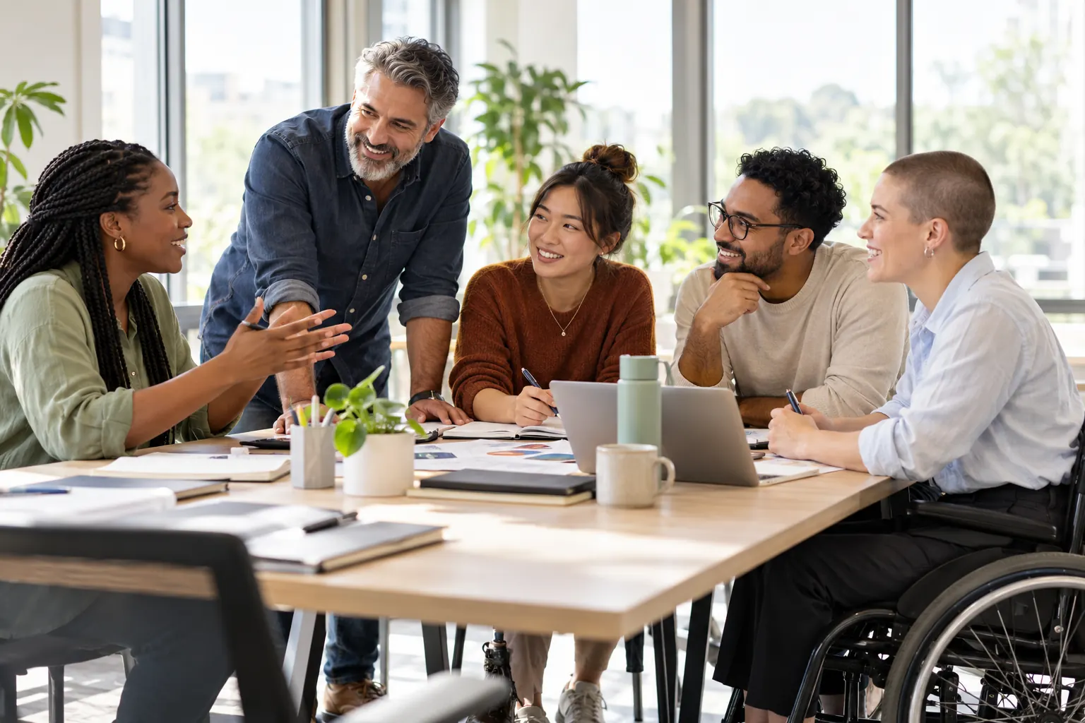 Coworkers of different ages and backgrounds, one using a wheelchair, work together around a table in a bright office.