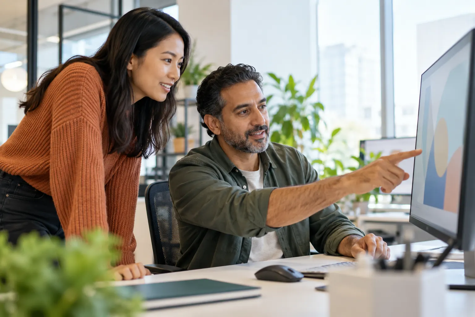 A young woman leans in to watch as an experienced colleague points at a chart on his monitor.