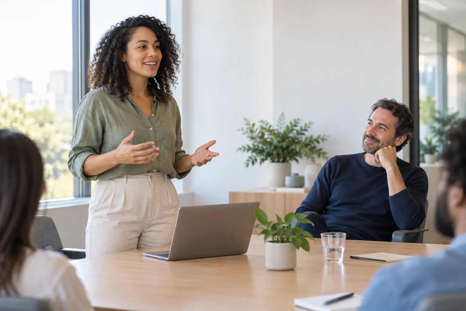 A smiling employee stands to present at a meeting table while her manager sits back and listens.