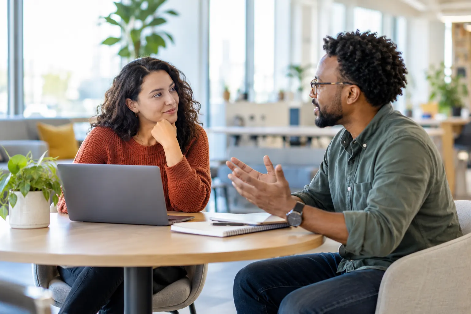 A woman listens with her chin on her hand while a colleague explains something across a round office table.