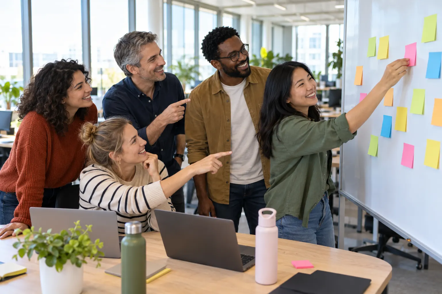 Five coworkers gather at a whiteboard covered in sticky notes while one adds another note.